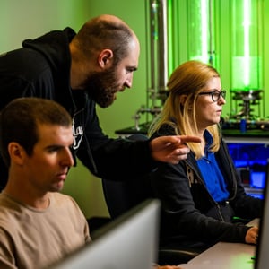 Photograph shows three people gathered around a computer screen. Two are sitting, one is standing behind them.
