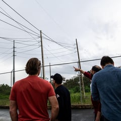 Photo shows several people looking at transmission lines next to a chain-link fence, behind which is lush vegetation.