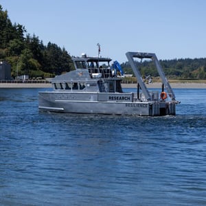 Photograph shows the RV Resilience vessel, a 50-foot-long silver boat, in Sequim Bay, with buildings of the PNNL Sequim campus in the background.