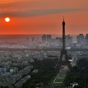 The glowing orange Sun shines over a lightly hazy Paris, the Eifel Tower standing, just right of center-frame.